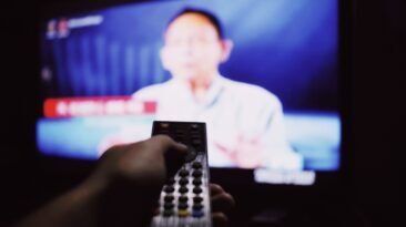 A person points a remote control toward a television in a dim living room.